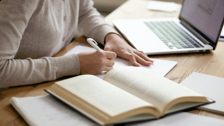 Person writing essay on paper with book and laptop open